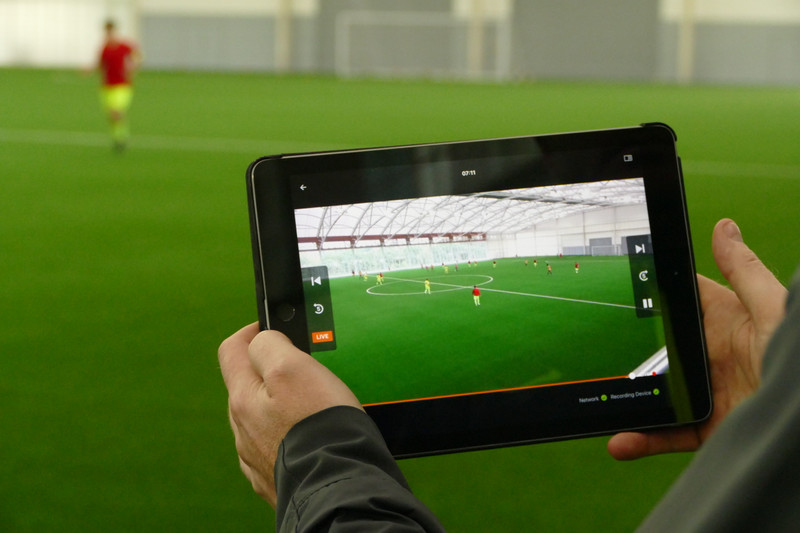 Homem reproduz vídeo da partida de futebol no tablet na área técnica do campo