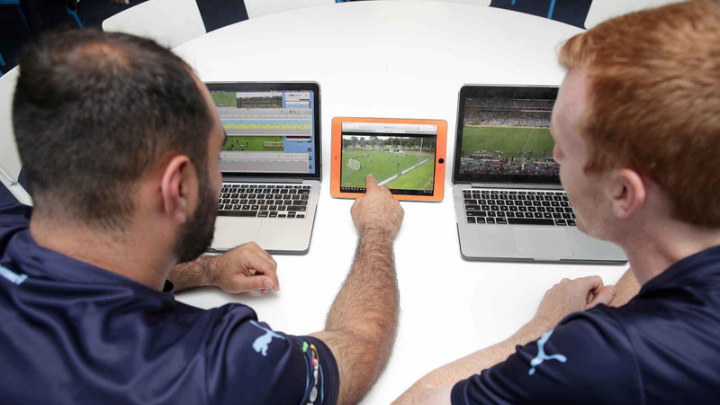 Dois jogadores do Sydney FC assistem a uma análise de partidas de futebol em dois laptops e um tablet